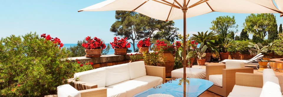 En hyggelig terrasse med hvide sofaer, en parasol, frodige planter og blomsterkrukker under den blå himmel.