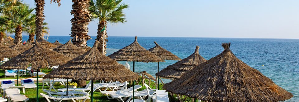 En strandlinje med parasoller af strå og palmer. Liggestole står klar til brug med udsigt over havet.