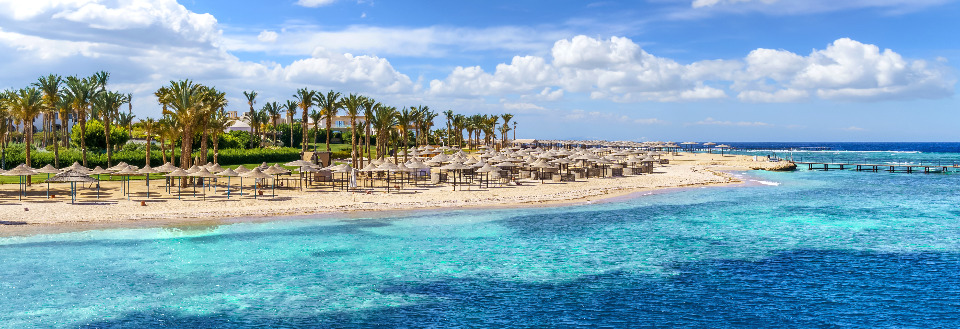 En tropisk strandpromenade med parasoller, palmer og en turkisblå hav foran under en klar himmel.