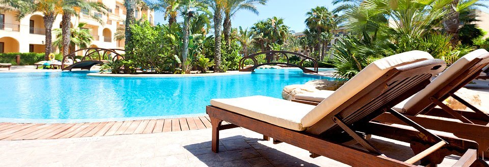 Et luksuriøst resort med en swimmingpool omgivet af palmer og liggestole under en solrig himmel.