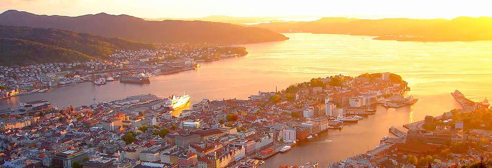 Billede af en by ved solnedgang med bjerge i baggrunden og en glitrende flod eller fjord foran.