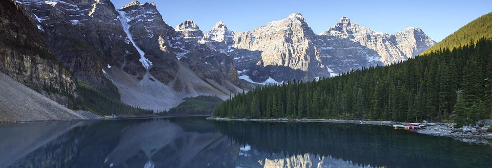 Billedet viser en bjergkæde reflekteret i en stille sø med en klar blå himmel og små både ved bredden.