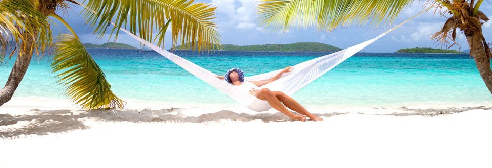 En person slapper af i en hængekøje mellem palmer på en tropisk strand med turkisblåt vand.