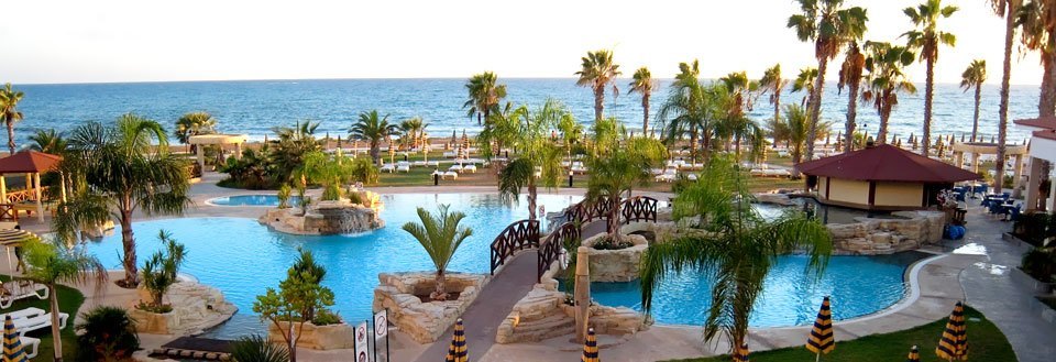 Swimmingpool på resort, omgivet af palmetræer med udsigt til havet ved solnedgang.
