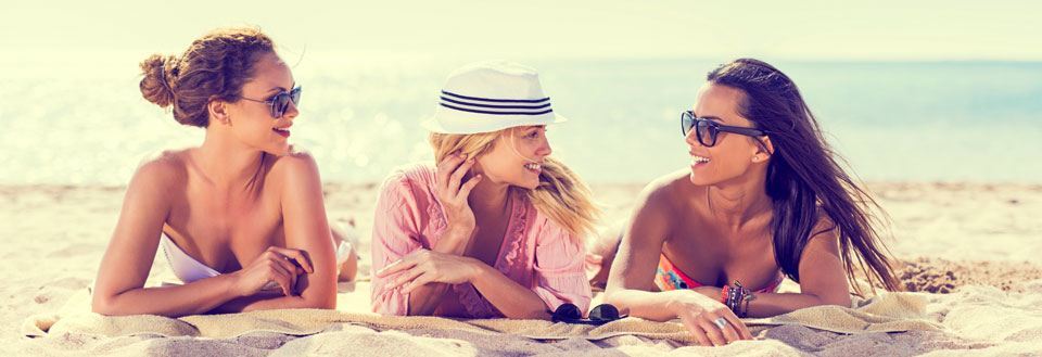 Tre kvinder smiler og snakker sammen på en solrig strand med blå himmel og hav i baggrunden.