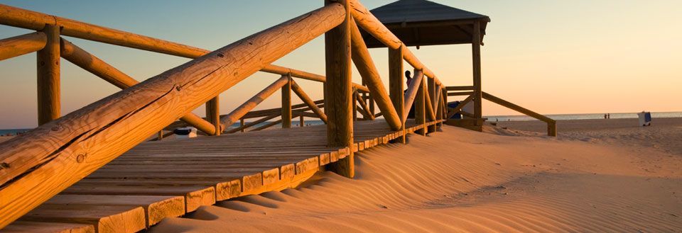 En træbro over sandet, der fører til et strandpavillon ved solnedgang.
