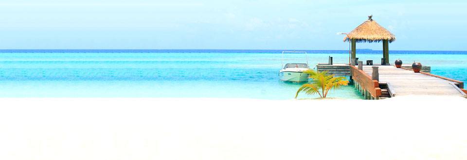 Hvid sandstrand med badebro, der fører ud til en stråparasol og en båd ved det turkisblå hav.