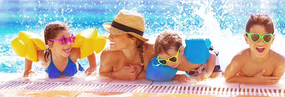 Børn med svømmevinger og farverige solbriller leger ved poolkanten i solskin.