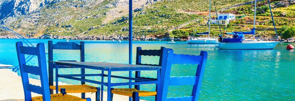 Blå stole foran en smuk bugt med klart vand og sejlbåde, omgivet af et kuperet landskab.