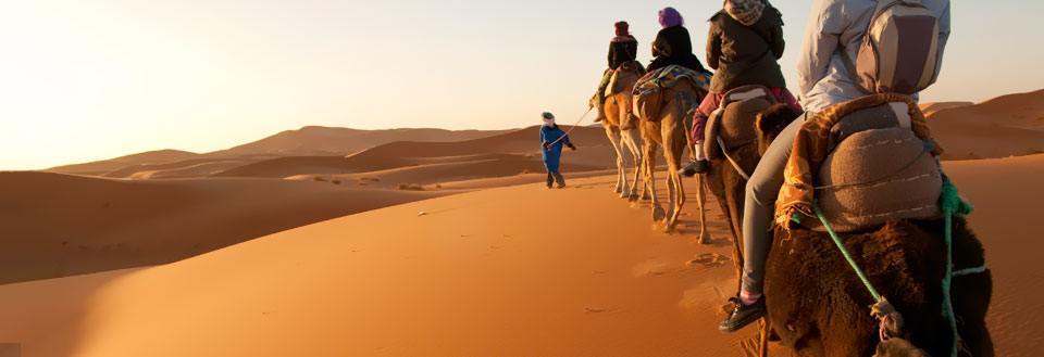 En gruppe mennesker rider på kameler gennem en ørken ved solnedgang, med sandklitter i baggrunden.