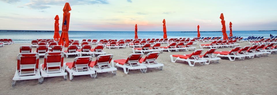 En tom strand med rækker af røde og hvide solstole og lukkede parasoller ved daggry.