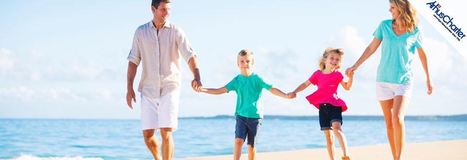 En familie på fire går hånd i hånd langs en solrig strand med blå himmel.