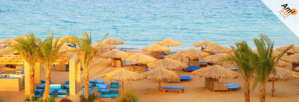 Strandscene med parasoller, strandstole og palmer ved kanten af klart blåt vand under en solrig himmel.