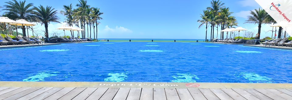 Et luksuriøst resortbassin med klart blåt vand, omgivet af solsenge og palmer med havudsigt.
