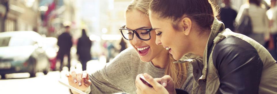 To kvinder smiler og ser på en smartphone sammen i en travl bygade.